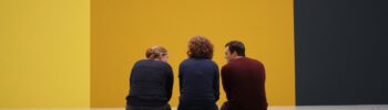 Drei Personen sitzen auf einer lehnenlosen Bank und betrachten eine bunte Wand.
